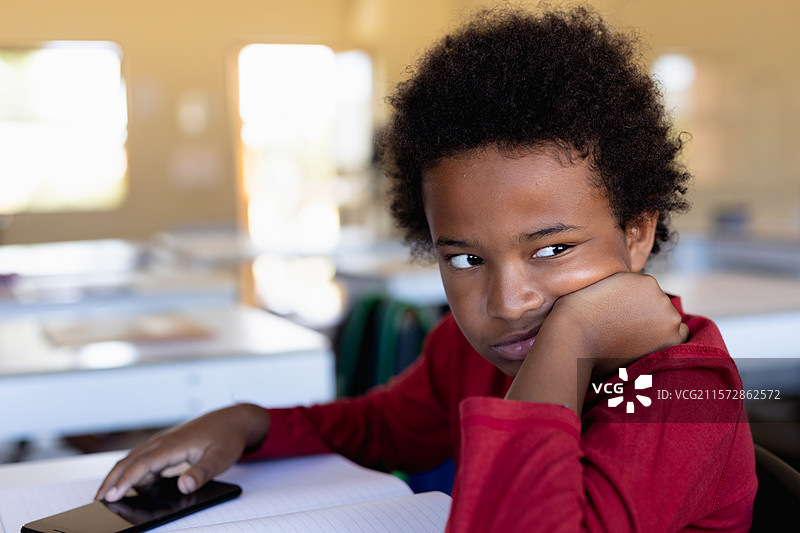 非裔美国男孩坐在教室的桌子旁，手里拿着笔记本和智能手机，目光向左凝视。图片素材