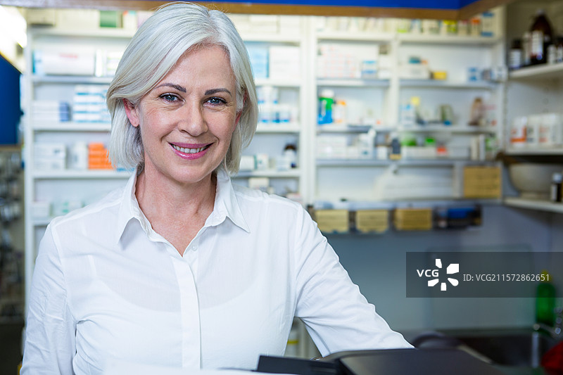 成熟的女性药剂师站在药房柜台前整理药品盒，面带微笑图片素材