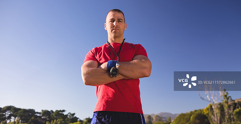 男性健身教练穿着运动服，双臂交叉站在场地上，手腕缠绕着护腕，手中拿着秒表。图片素材