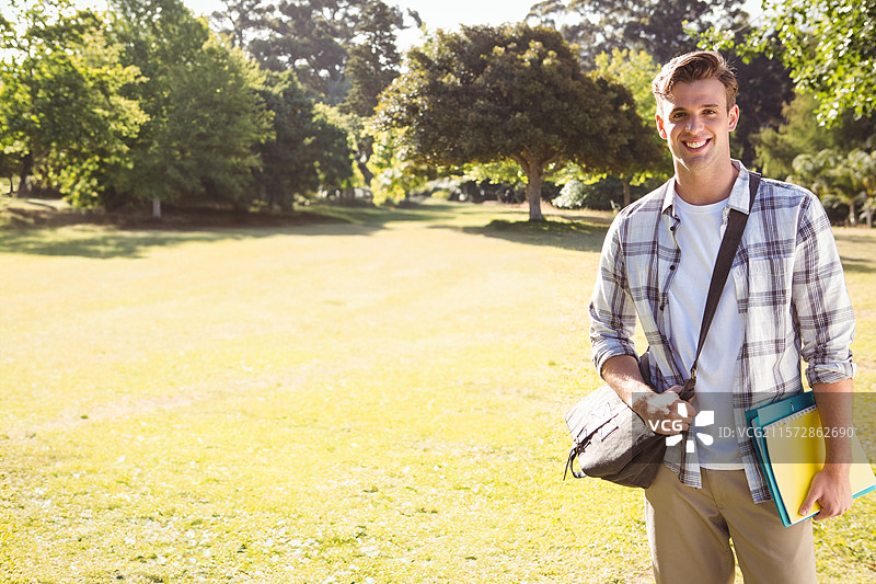 男学生站在阳光明媚的校园草坪上，手里拿着五彩缤纷的笔记本和邮差包，留有空白空间。图片素材