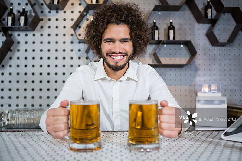 男人手持两杯泡沫啤酒，靠在带有蜂窝架的打孔酒吧柜台后面。图片素材