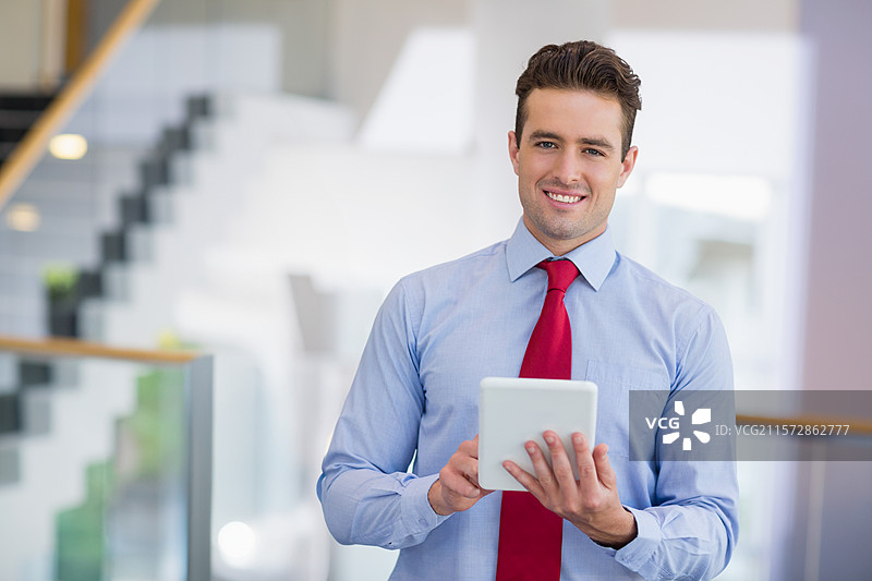 男士在办公室大堂站着，手持平板电脑，穿着衬衫和领带，靠近玻璃栏杆。图片素材