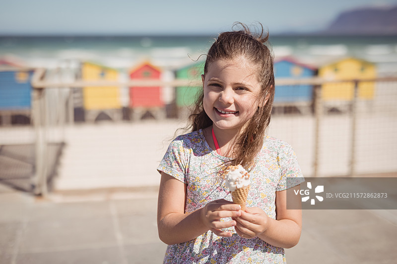 微少女微笑着，手里拿着撒有糖屑的冰淇淋蛋筒，站在海滩步道的栏杆旁，背景是五彩缤纷的海滩小屋。图片素材