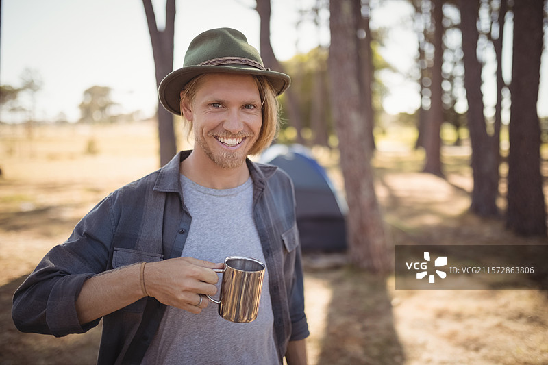 穿着格子衬衫、戴着绿色毛毡帽的男性露营者，在松树林营地中手持金属露营杯。图片素材