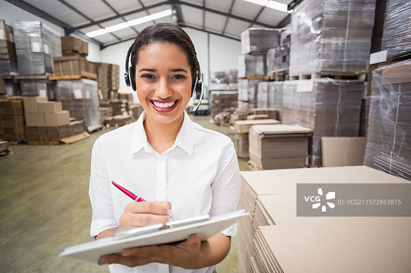 非裔美国女性在仓库检查箱子，手持夹板和红色圆珠笔，佩戴耳机。图片素材