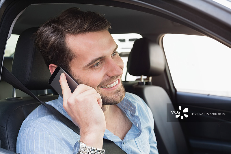 微笑的男子穿着蓝色衬衫，手持智能手机靠在耳边，同时在轿车内系安全带。图片素材