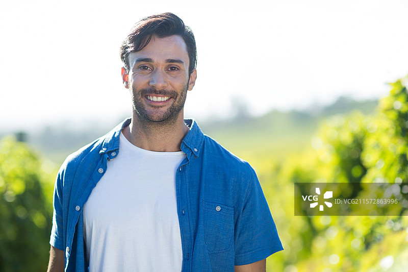 站在阳光明媚的葡萄藤间，微笑着看向镜头，穿着蓝色短袖衬衫。图片素材