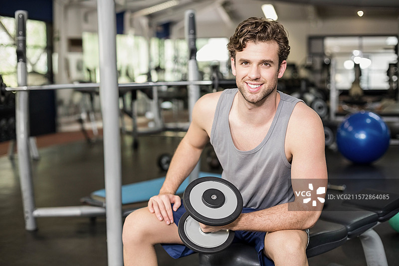 健身房里，坐在举重凳上的健壮男子举着哑铃，旁边有深蹲架和稳定球。图片素材