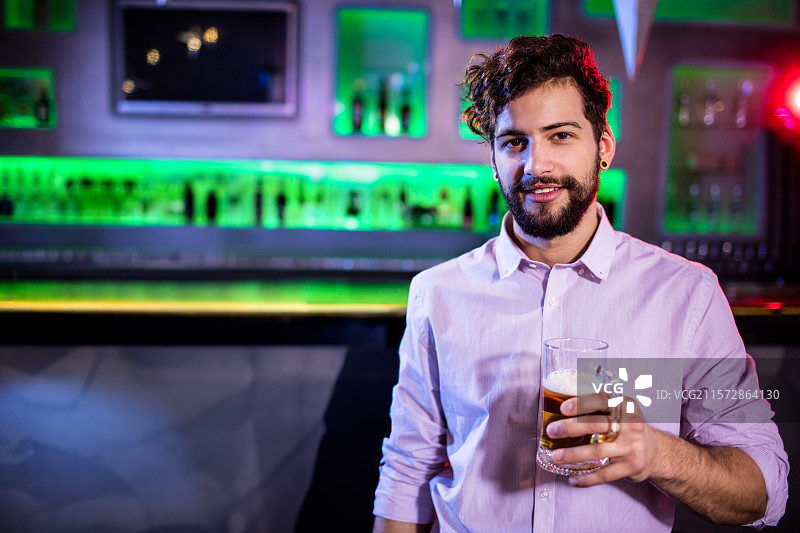 在酒吧柜台前，留着胡子的男人手持啤酒杯，背后是装满瓶子的霓虹灯架，留有空白空间。图片素材