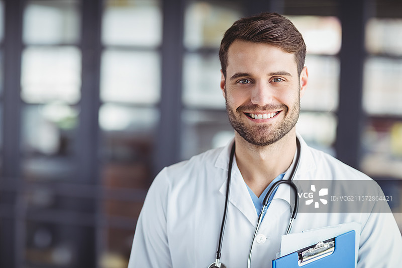 医生在诊所接待区站着微笑，手持夹板，佩戴听诊器。图片素材