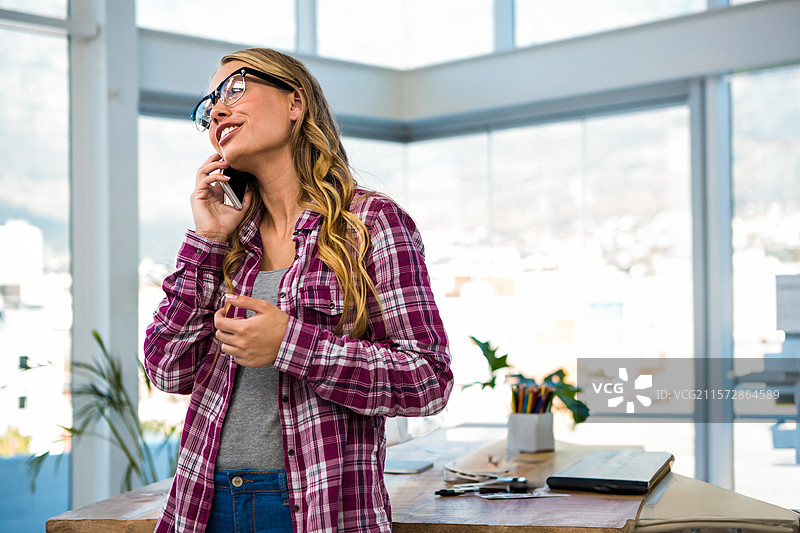 白人女性专业人士在办公室的桌子上用智能手机通话，窗边有一盆植物。图片素材