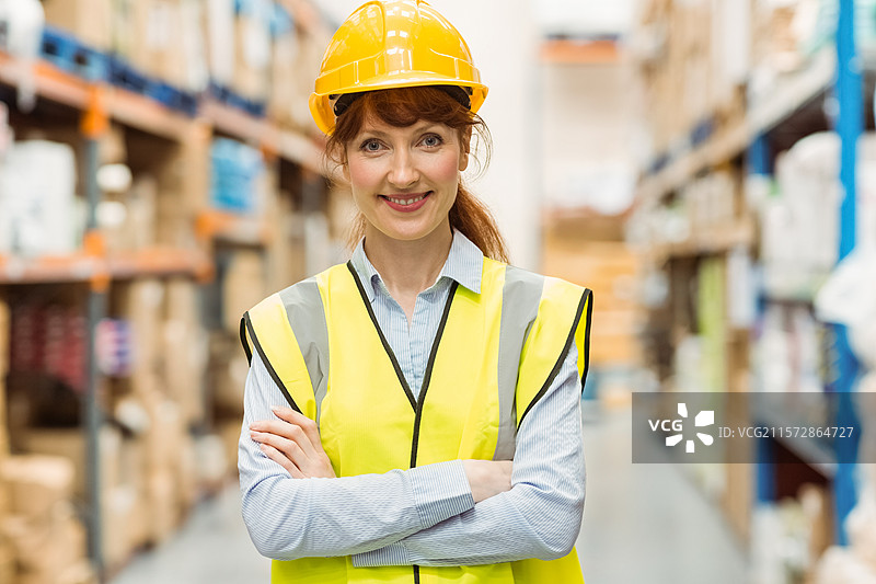 中年女性站在仓库过道中，双臂交叉，戴着安全帽和安全背心。图片素材