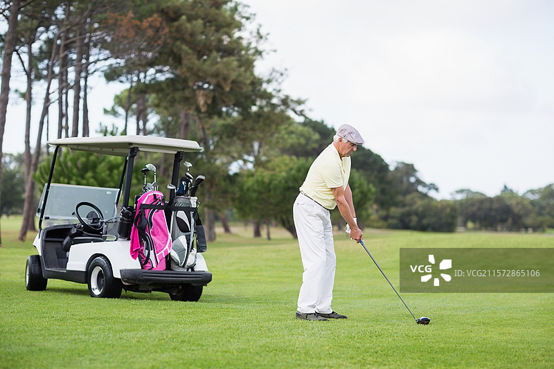 一位资深高尔夫球手正在球道上准备挥杆，旁边有一辆高尔夫球车，车上放着粉色和黑色的球包。图片素材