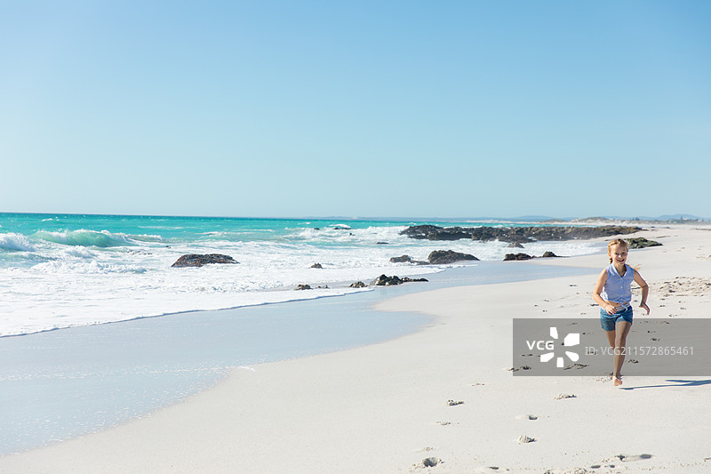 赤脚的小女孩在沙滩上奔跑，身旁是碧蓝的海浪和黑色的岩石，留有空白空间。图片素材