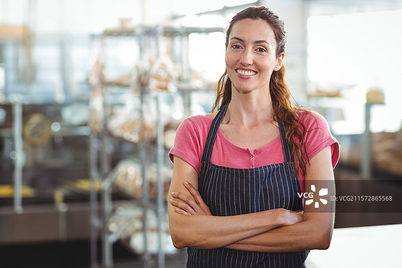 女性面包师站在面包店的柜台后，穿着条纹围裙，微笑着，身后是面包架。图片素材