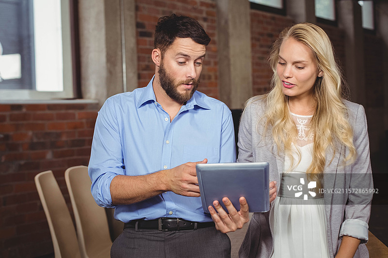 身穿商务服装的同事在现代办公室使用平板电脑，周围有堆叠的椅子和桌子。图片素材