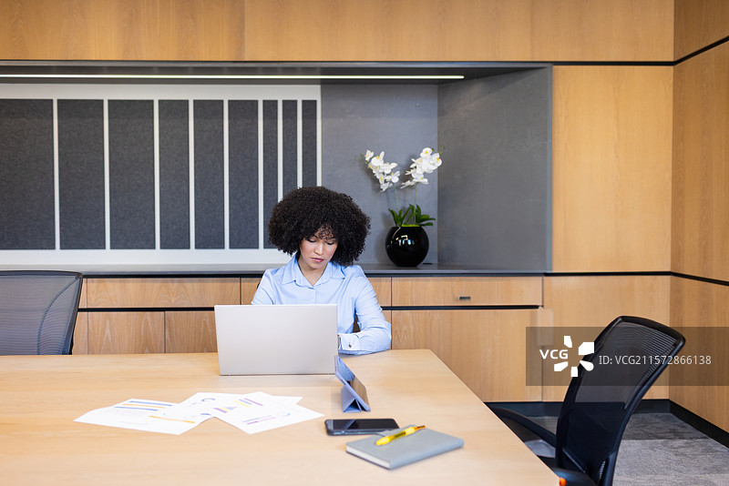 一位穿着蓝色衬衫的非裔美国女性坐在会议桌前，正在用笔记本电脑输入数据，桌上有图表。图片素材