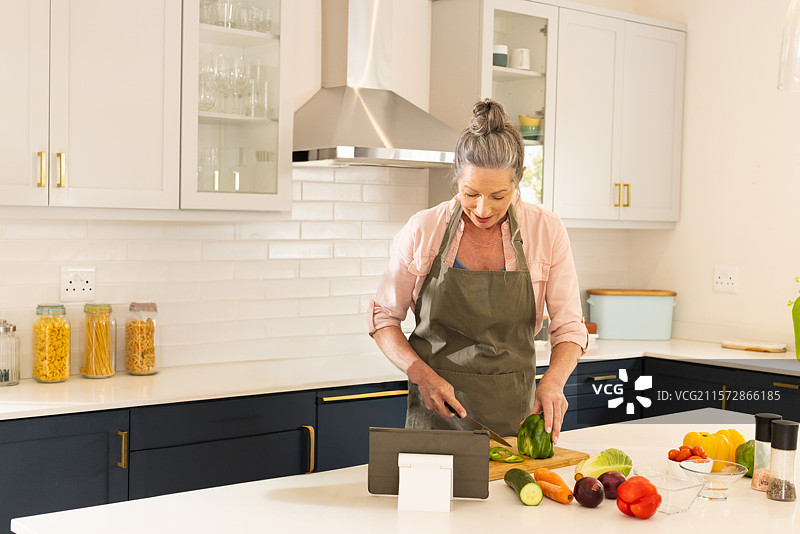 家庭厨师在厨房的白色石英台面上切割绿色甜椒，旁边有一个平板电脑支架。图片素材