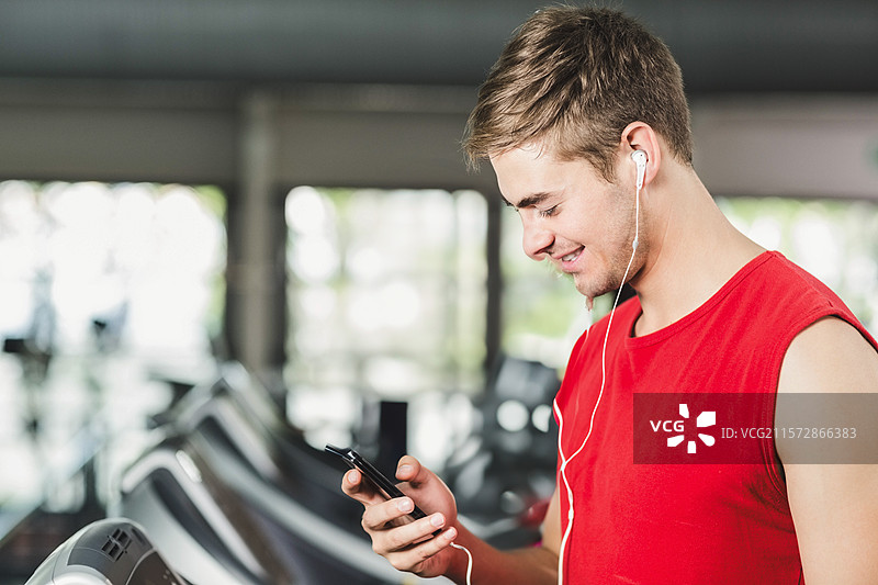 穿着红色运动服的男子在健身房的跑步机旁使用智能手机，耳机在耳边，有空白空间。图片素材