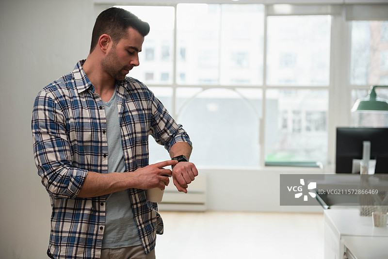 男子在现代办公室的大窗户旁查看智能手表，旁边有显示器和绿色台灯，留有空白空间。图片素材