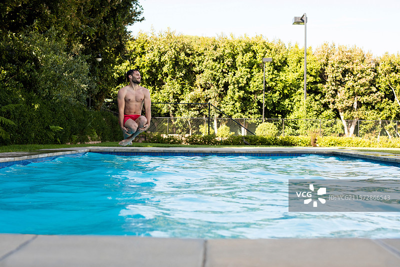 穿着红色泳裤的健壮男子蹲在混凝土泳池边缘，准备跳入蓝色水中图片素材