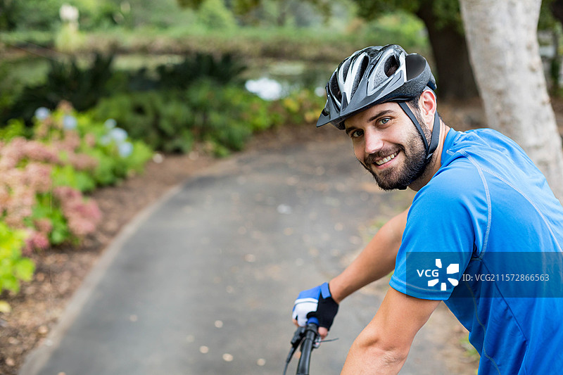 骑自行车的男士微笑着回头，佩戴着头盔和手套，骑在铺砌的小路上，留有空白空间。图片素材