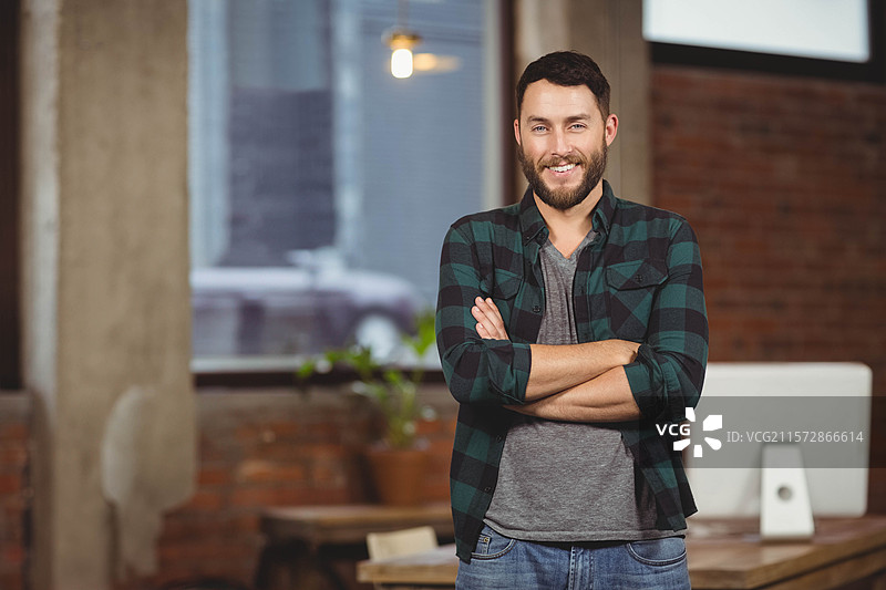 中年男子双臂交叉站在工业窗旁的阁楼办公室里，旁边有显示器，留有空白空间。图片素材