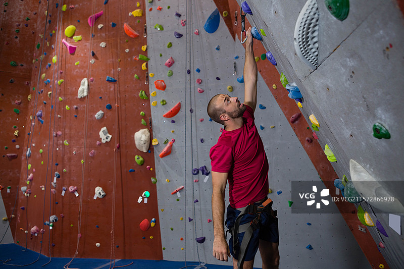 攀岩者在健身房内攀爬墙壁，使用安全带和绳索，脚下是铺有彩色把手的垫子。图片素材