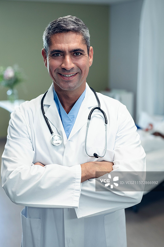 穿着白大褂和听诊器的老年男性医生站在医院病房内，旁边是病床。图片素材