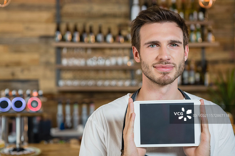 男调酒师手持平板电脑，在酒吧柜台前展示空白屏幕，周围有啤酒龙头和酒瓶。图片素材