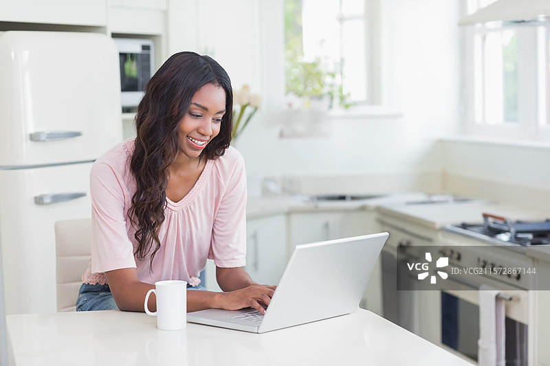 非裔美国女性在厨房台面上微笑着使用笔记本电脑，旁边有一个杯子和花。图片素材
