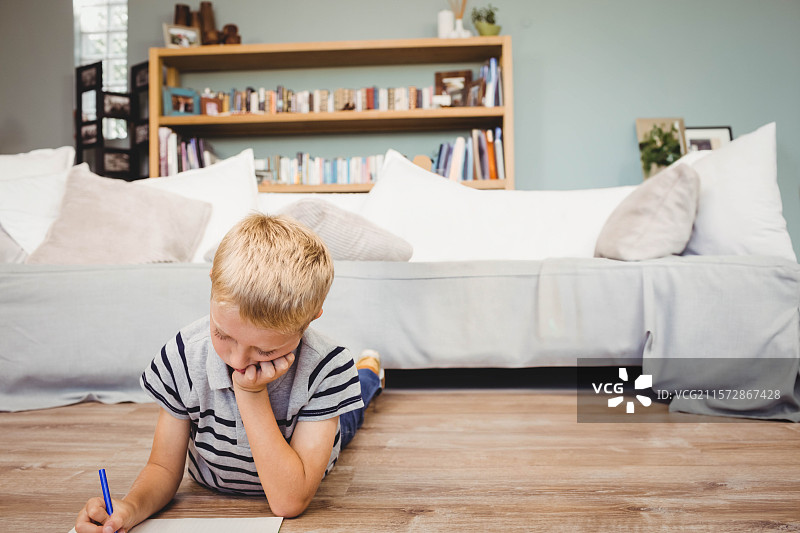 孩子在灰色沙发旁的木地板上写字，手里拿着蓝色的笔，身后是书架。图片素材
