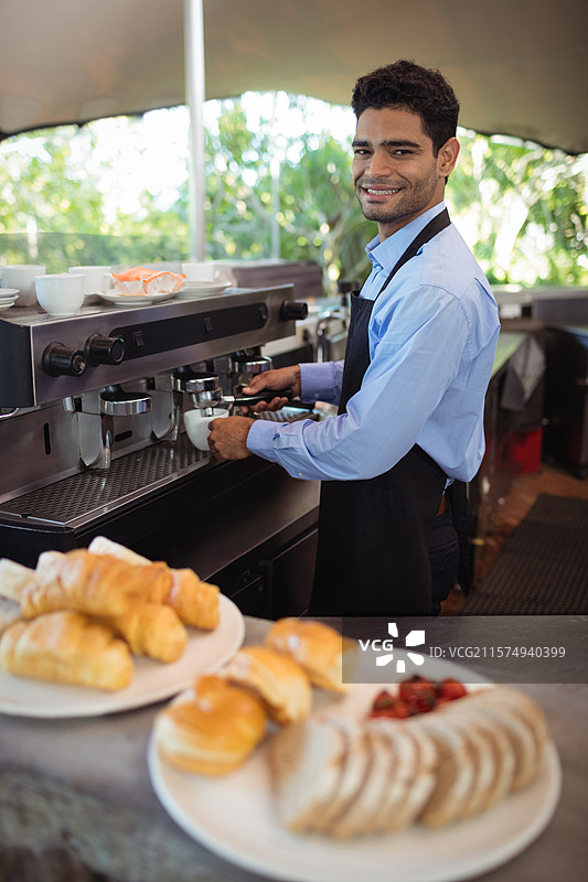 在户外咖啡馆柜台后操作浓缩咖啡机的男士，旁边有糕点盘和杯子，留白空间图片素材