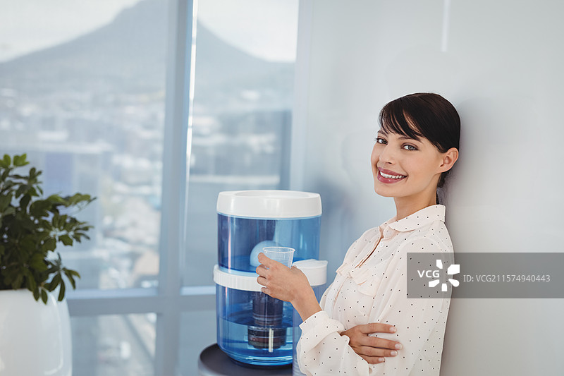 办公室女性站在蓝色饮水机旁，手持杯子，窗边有盆栽植物，留有空白空间。图片素材
