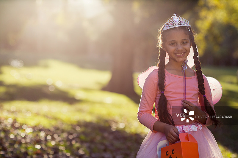 南瓜形状的万圣节糖果袋坐在阳光洒落的树下的草地上，周围散落着树叶。图片素材