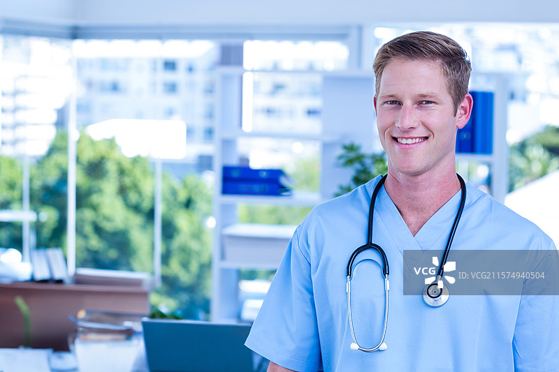 穿着蓝色手术服和听诊器的医生站在接待台前，手里拿着笔记本电脑和文件。图片素材