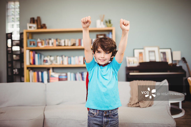 男孩在家中的客厅里站着，举起双臂，穿着红色斗篷，靠近沙发和钢琴。图片素材