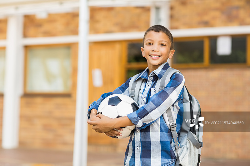 男孩站在砖砌的学校建筑前，手里拿着足球，背着书包。图片素材