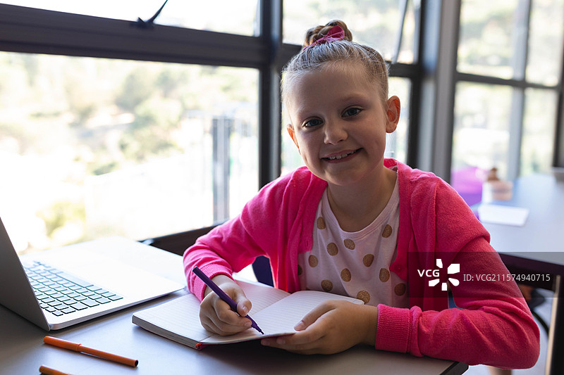 微笑的女孩在窗边的桌子上用紫色钢笔在笔记本上写字，旁边有一台笔记本电脑。图片素材