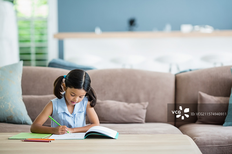 孩子在客厅的木质咖啡桌上用彩色铅笔写字，旁边放着一本打开的笔记本。图片素材