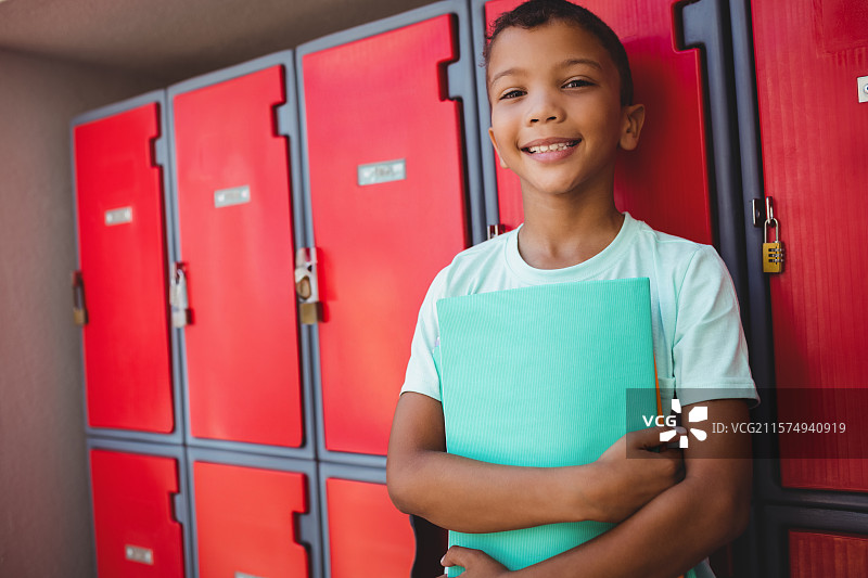 男孩站在明亮的红色金属储物柜旁，胸前抱着青绿色的活页夹，在学校走廊里。图片素材