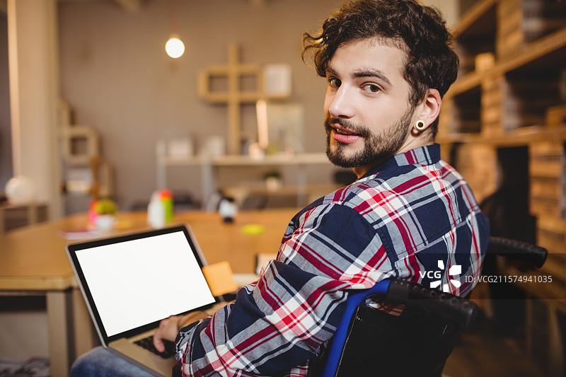 坐在办公桌旁的胡子男人，坐在轮椅上用笔记本电脑打字，周围有书架和箱子。图片素材