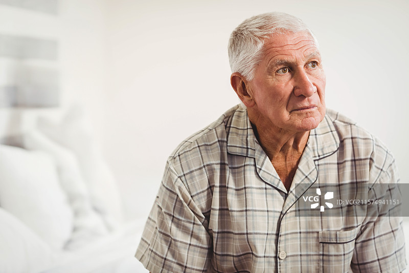 老年男子坐在床边，穿着格子睡衣上衣，柔和的灯光照耀着床头的卧室。图片素材
