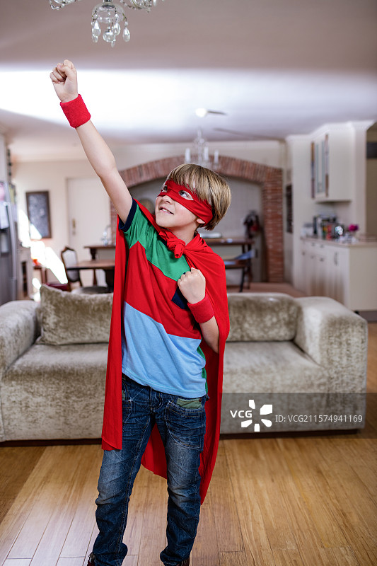 孩子在客厅里英勇地摆姿势，身穿红色斗篷和面具，旁边是水晶吊灯。图片素材