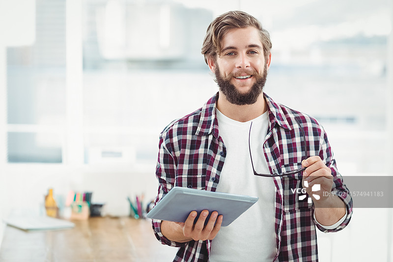 留着胡子的男人在现代办公室里拿着平板电脑和眼镜，木桌上放着笔，窗外是城市天际线。图片素材