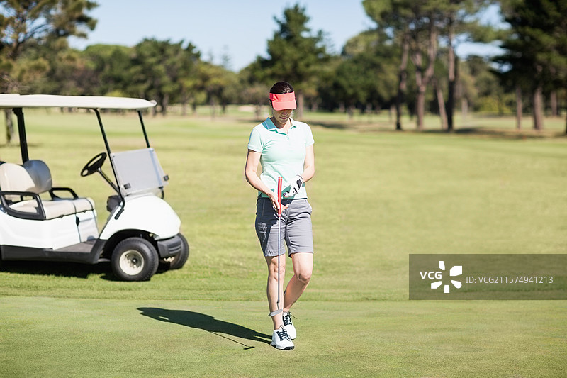 女性高尔夫球手站在果岭上，手持推杆和高尔夫球，旁边有一辆白色高尔夫球车。图片素材
