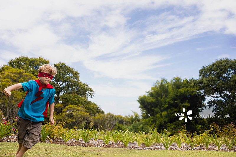 穿着蓝色T恤、展示红色闪电标志、红色斗篷和眼罩的小男孩在草坪上奔跑图片素材