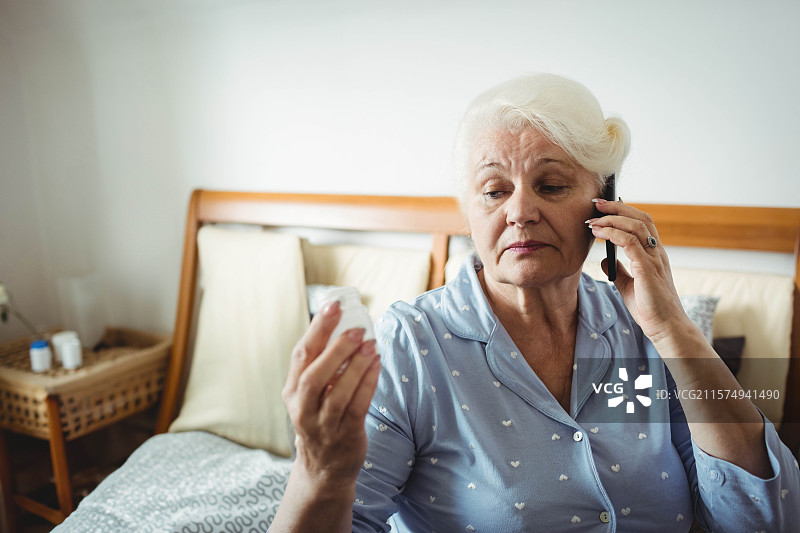 一位老年女性坐在卧室的床上，阅读药瓶并拿着智能手机，周围有空白空间。图片素材