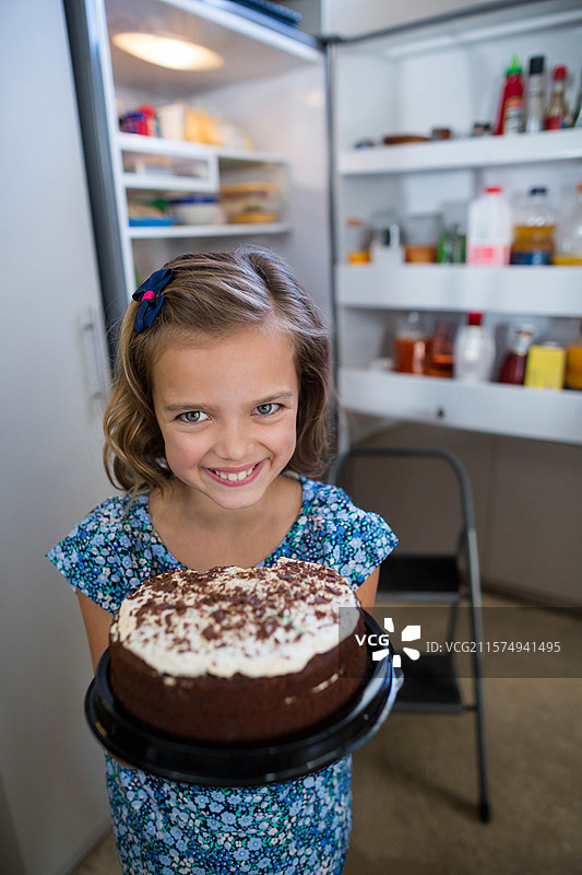 穿着蓝色裙子的微笑女孩在厨房的开放冰箱旁拿着一块白色糖霜巧克力蛋糕图片素材