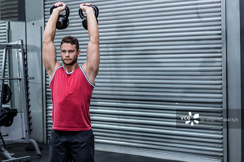 穿着红色衬衫的男性运动员在健身房举起壶铃，背景是重物架和金属卷帘门。图片素材